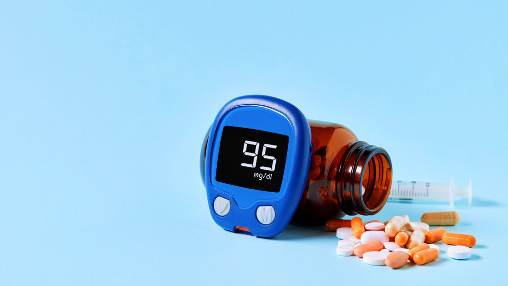 Glucose meter displaying ideal blood sugar range, colorful medical pills spilling out of drug bottle and a syringe on blue background with copy space. Diabetes and healthcare concept.