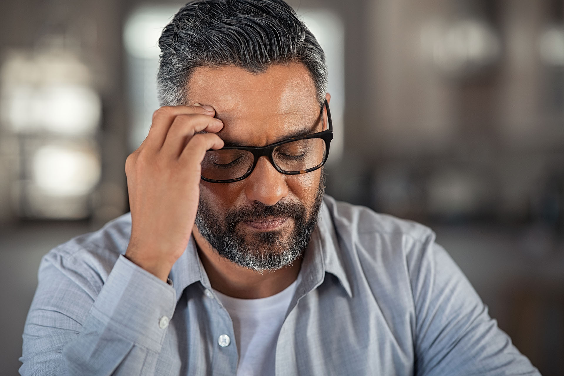 Frustrated middle aged man with hand on head sitting on couch at home. Close up face of stressed indian businessman wearing eyeglasses with eyes closed. Worried middle easter business man with terrible migraine, overload and overworked concept.